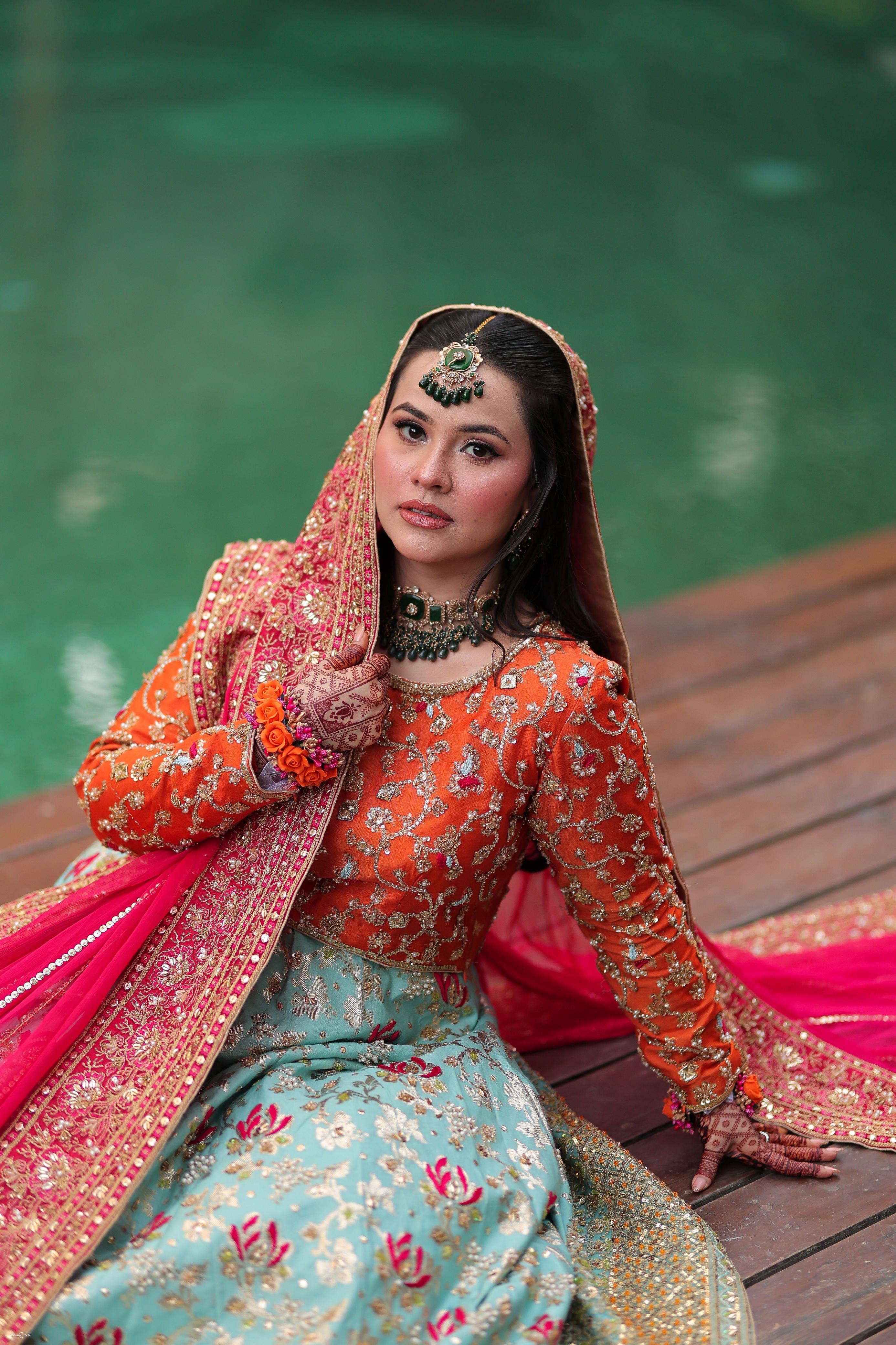 Bride in orange and gold bridal couture with pink dupatta and aqua skirt by the water
